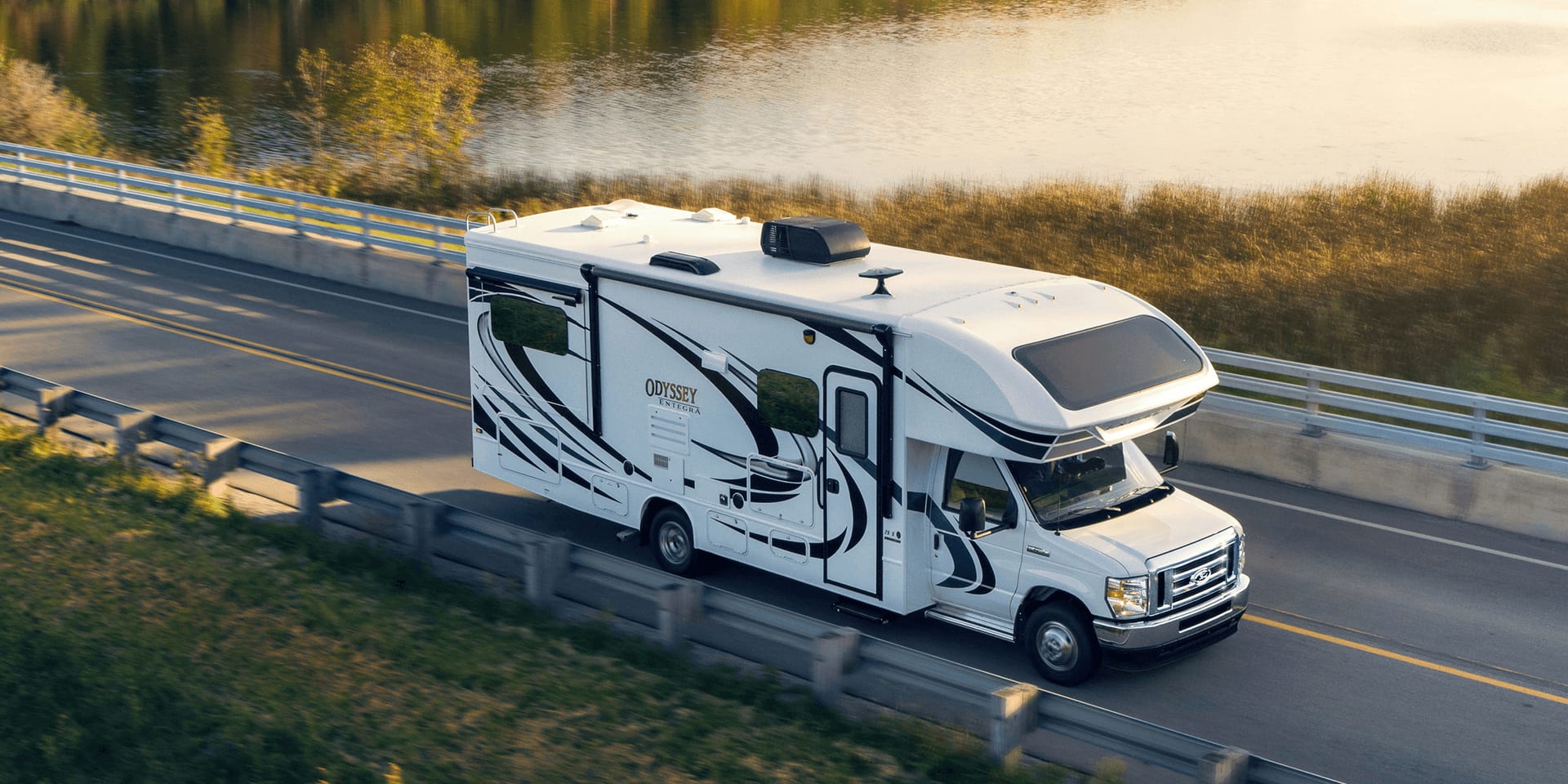 A Class C motorhome is driving past a body of water.