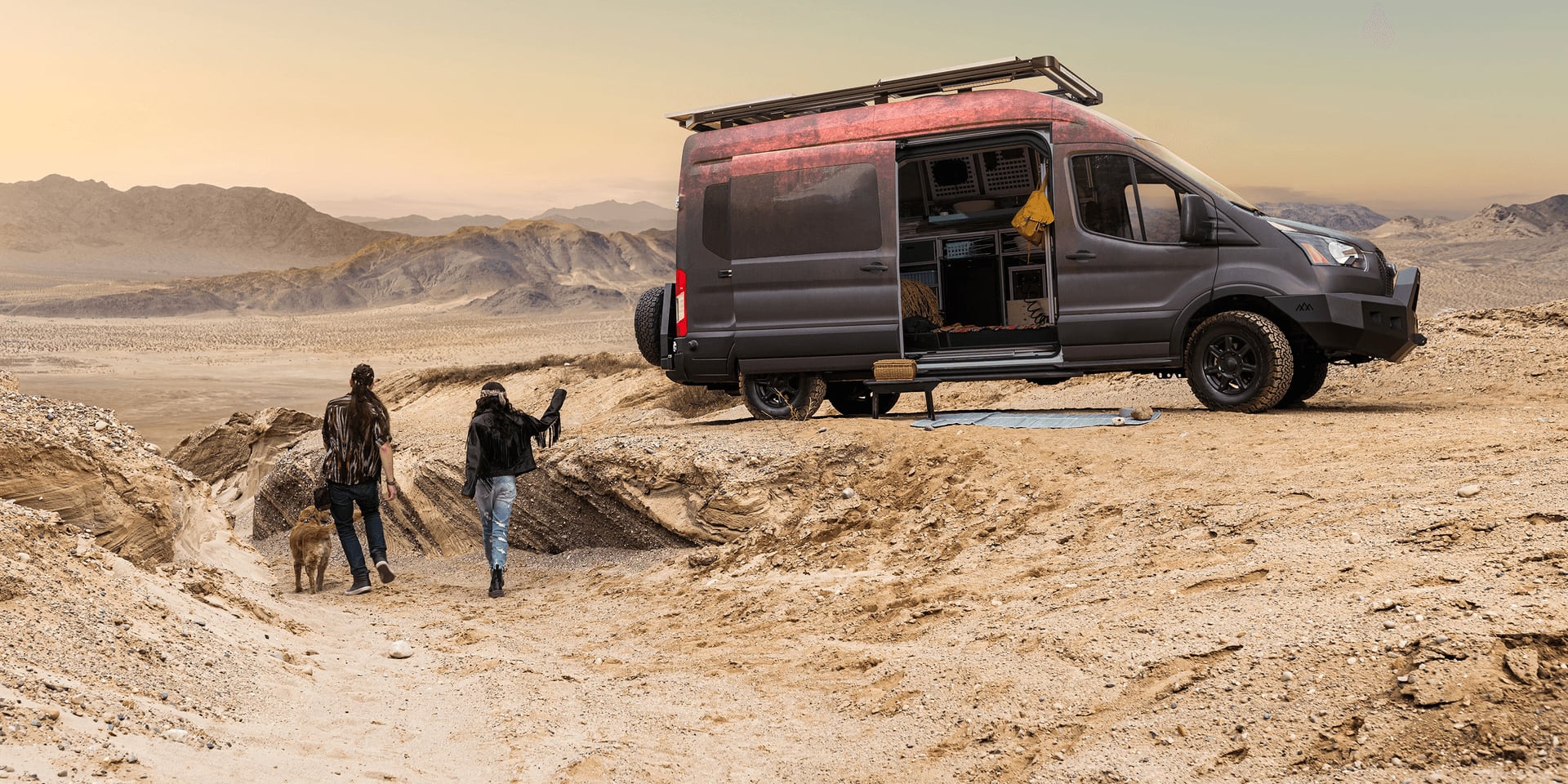 A man is on one knee in front of a red Transit campervan in the desert, taking a picture of a woman crouched next to a golden retriever.